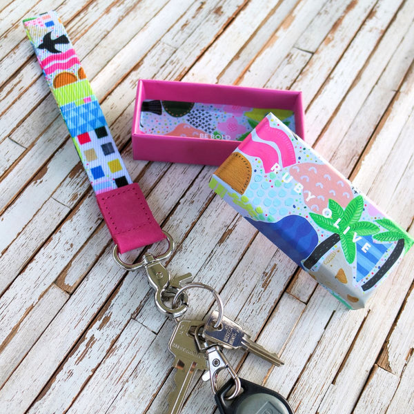 A colorful patterned wristlet keychain with attached metal key ring and keys, shown on a whitewashed wooden surface alongside a pink rectangular box and a matching rolled pouch.