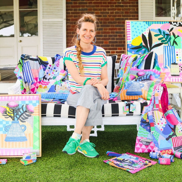 A woman sits on a white bench in front of a house porch, surrounded by colorful patchwork quilts, art prints, and fabric pieces. She is wearing a striped t-shirt and gray pants, with bright green sneakers.