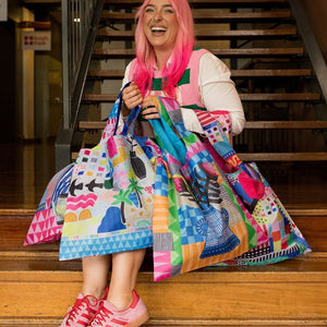 A woman with bright pink hair sits on wooden stairs holding a large, colorful patchwork tote bag. She is wearing a white and green striped top and pink sneakers.