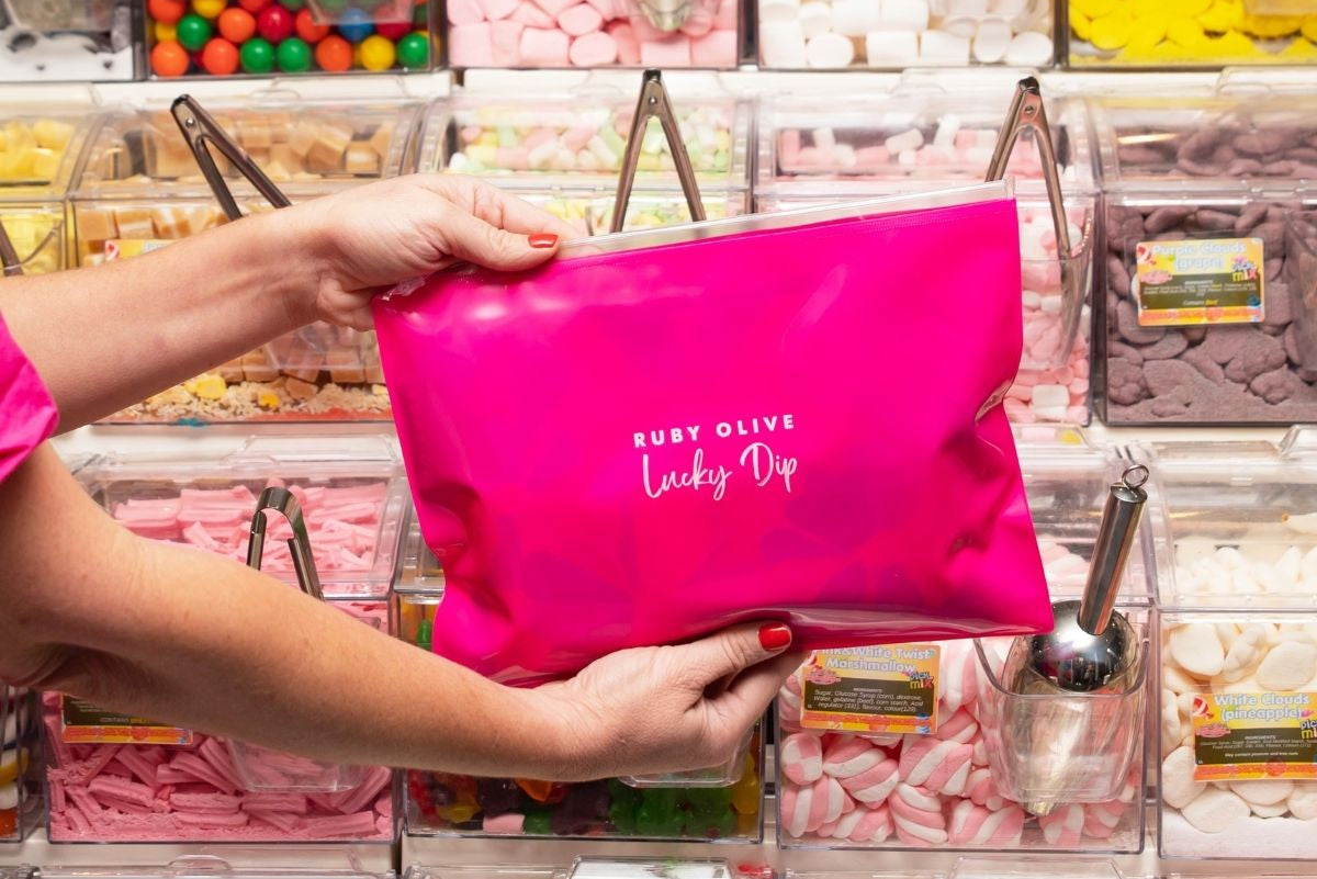 Person holding a pink pouch labeled 'Ruby Olive Lucky Dip' in front of a candy display.