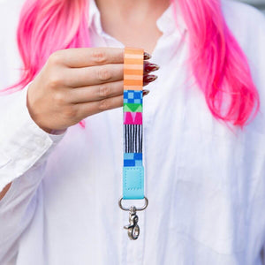 A person with bright pink hair holds up a colorful woven keychain strap with a light blue leather end and a silver lobster clasp. The person is wearing a white button-down shirt.