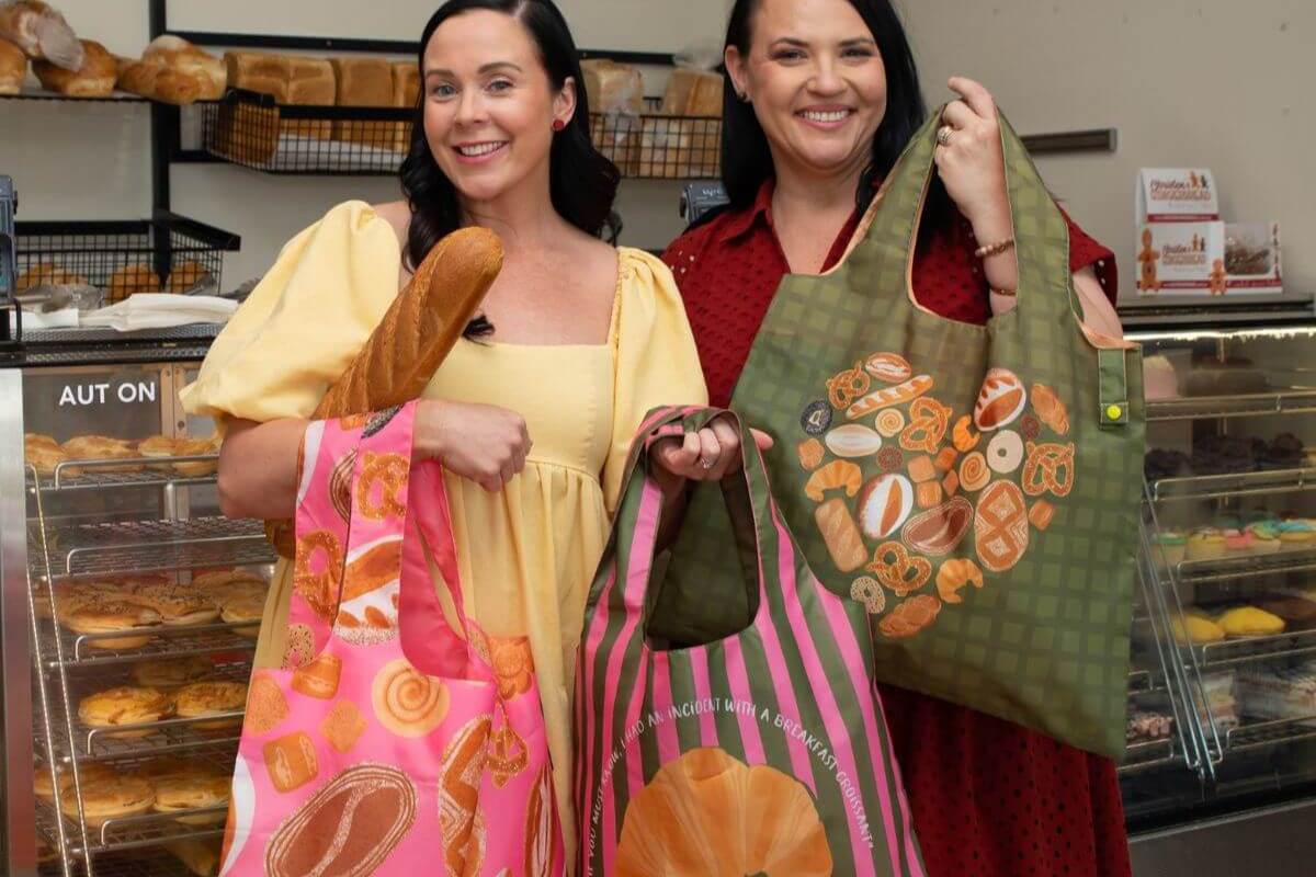 Two women holding colorful tote bags with bread designs in a bakery setting.