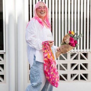 A woman with bright pink hair stands outdoors in front of a white slatted wall, holding a pink patterned shopping bag and a bouquet of flowers. She wears a white top, light blue jeans, and pink glasses.