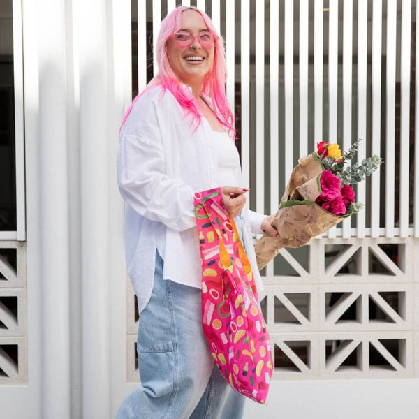 A woman with bright pink hair stands outdoors in front of a white slatted wall, holding a pink patterned shopping bag and a bouquet of flowers. She wears a white top, light blue jeans, and pink glasses.