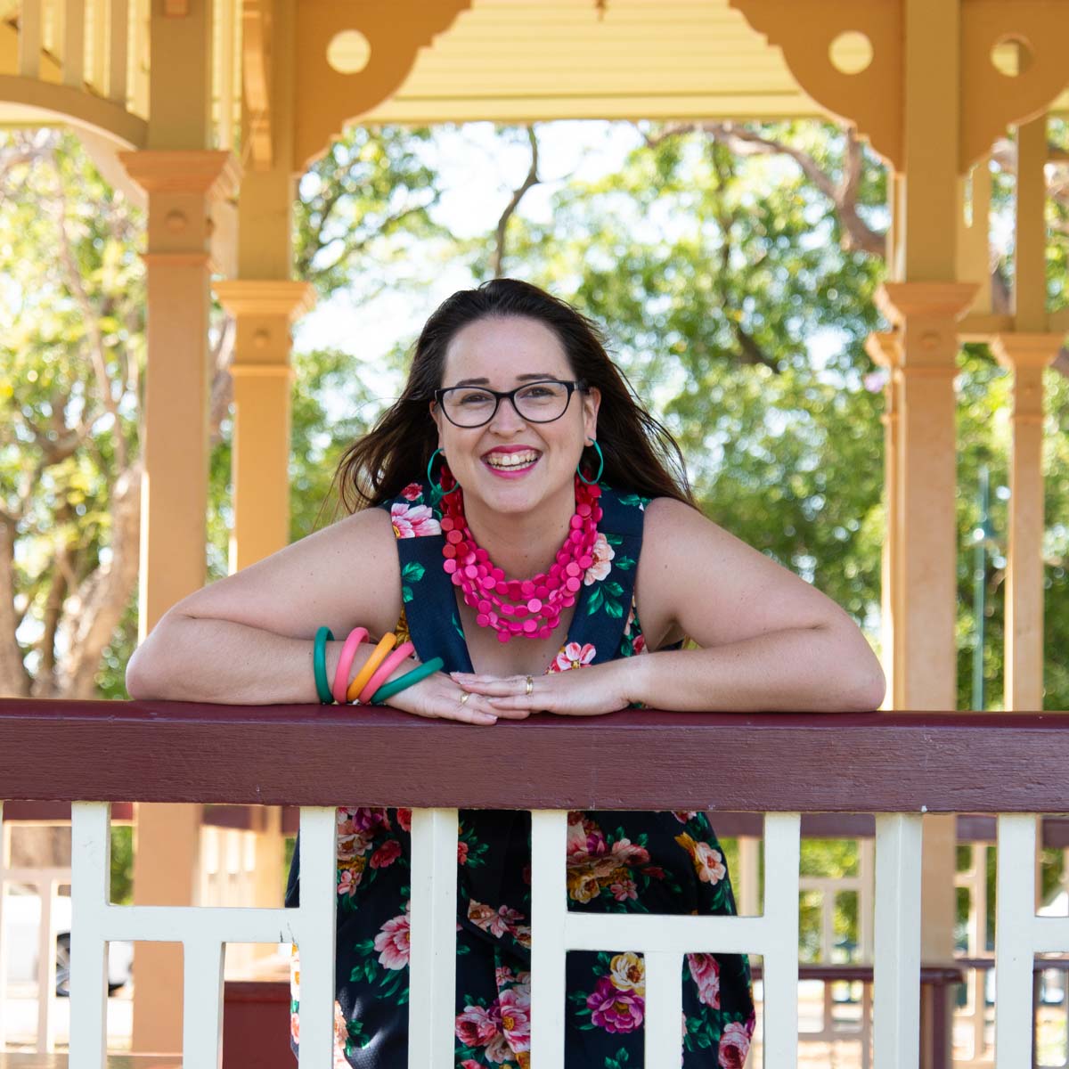 Happy dark haired women in a pergola wearing flower dress and bright multi coloured necklace.
