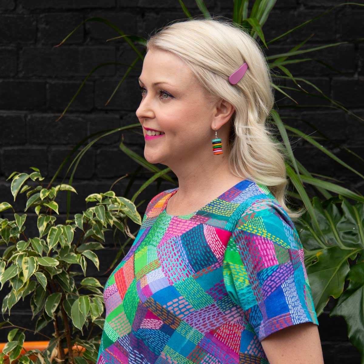 Side profile of blonde women wearing multi coloured stacked earrings and light pink clip.
