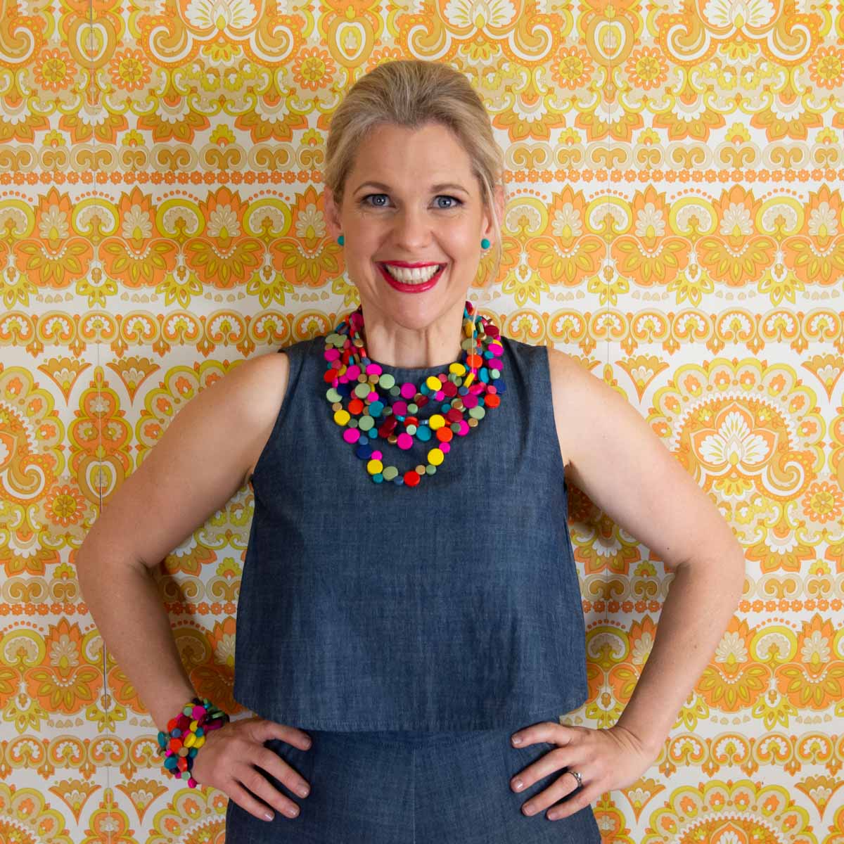 Smiling woman wearing stacked multi coloured necklace and bracelet in front of yellow and orange vintage wallpaper.